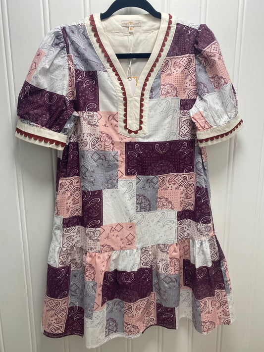 A short-sleeve patchwork dress hanging on a black hanger against a white paneled wall. The dress features a mix of purple, pink, gray, and white patterned fabric squares with a cream V-neckline and red trim. Simply Southern Dress.