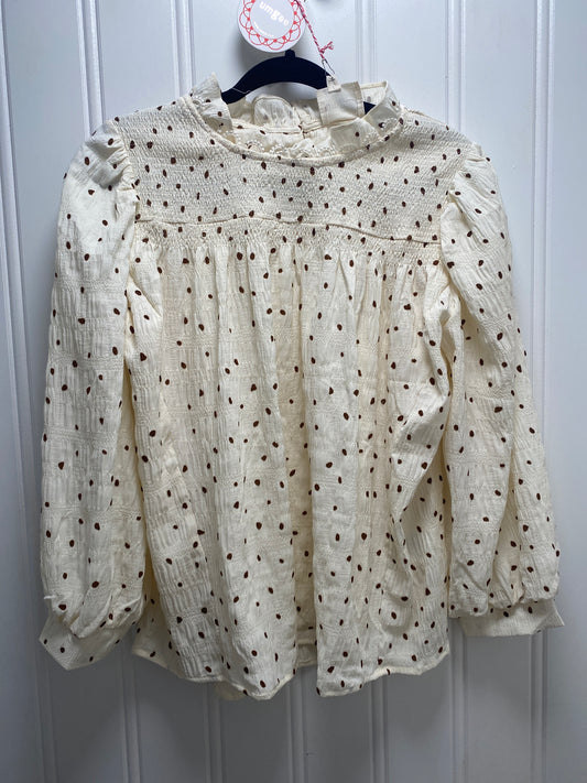 An off-white textured blouse with brown polka dots, featuring a smocked bodice and ruffled high neck, hanging on a black hanger against a white paneled door.