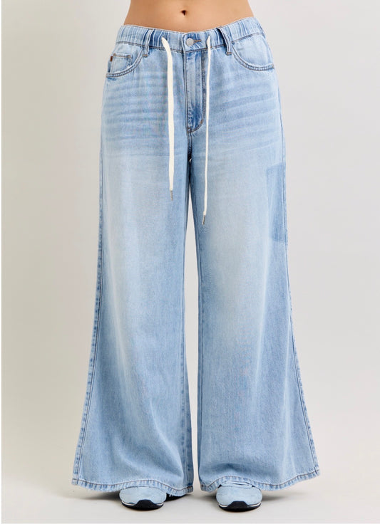 Light wash blue wide-leg denim jeans with a white drawstring waist, shown on a model from the waist down against a light gray studio background.  Judy Blue Palazzo pants