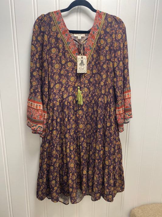 A bohemian-style long-sleeve dress with a dark purple base, yellow and orange floral print, and red patterned trim on the neckline and cuffs, hanging on a black hanger against a white paneled wall.  Dress by Easel