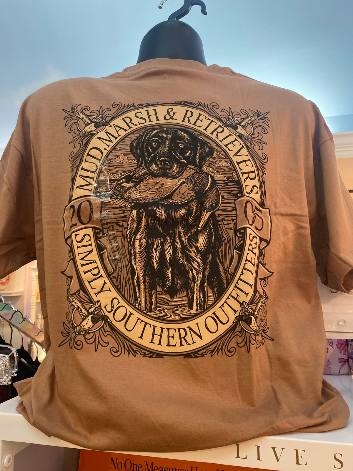 A tan graphic t-shirt displayed on a black hanger mannequin, showing the back with a large circular print of a dog holding a duck and the text 'MUD MARSH & RETRIEVERS SIMPLY SOUTHERN OUTLIERS'.