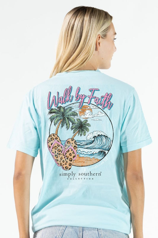 Woman wearing a light blue t-shirt with a palm tree and flip-flop design, featuring the text 'Walk by Faith' and 'Simply Southern Collection'.
