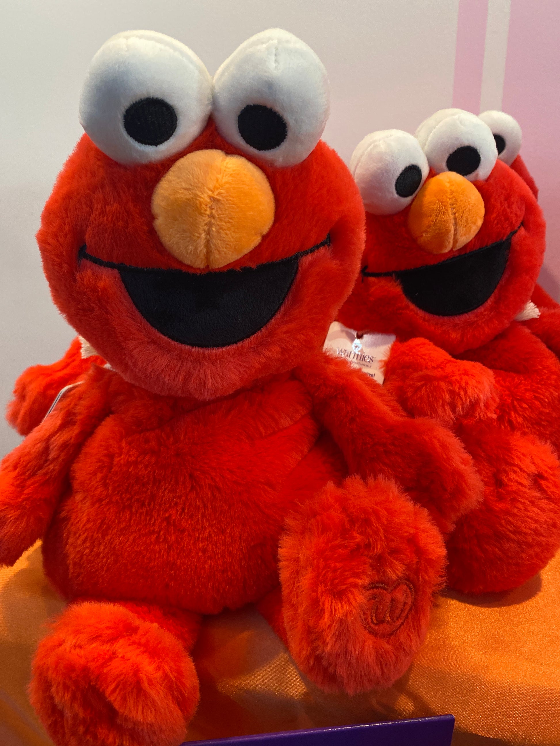 Red plush Elmo toys with large white eyes and orange noses, sitting on an orange surface against a light pink and white background.