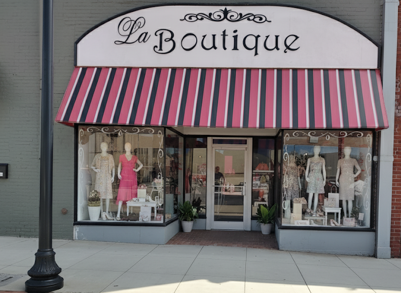 A storefront for 'La Boutique' with a red, black, and white striped awning, glass windows displaying mannequins in dresses, and a sidewalk in front.